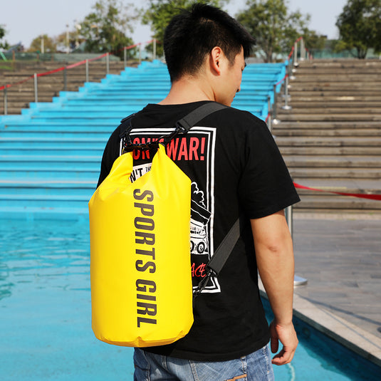 Person wearing a black t-shirt with a graphic design and carrying a yellow 'SPORTS GIRL' bag by a pool.