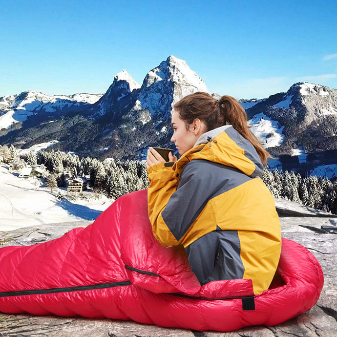 Person sitting in a red sleeping bag with snow-covered mountains in the background
