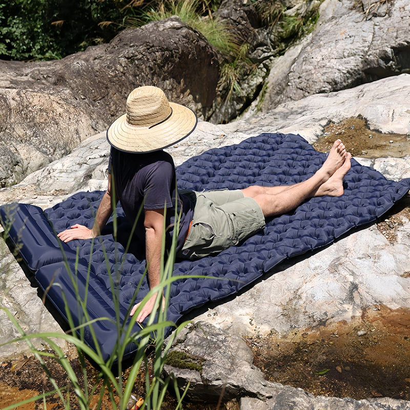 Load image into Gallery viewer, Person lying on a blue inflatable camping mat in a natural setting with rocks and water.

