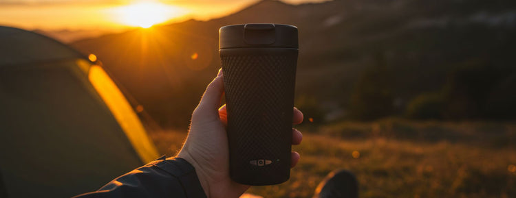 Hand holding a black travel mug with a scenic sunset background